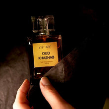 Load image into Gallery viewer, Oud Khashab — A rich woody unisex oud perfume — 50ml amber glass bottle. A  deep patchouli oud, sandalwood, leather &amp; smoky fragrance — EsQue Parfum — held in hand with soft dark fabric against warm moody background - vegan and oil based 